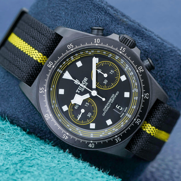 Tudor Black Bay Chronograph with black dial and yellow accents on fabric strap, resting on blue and green fabric